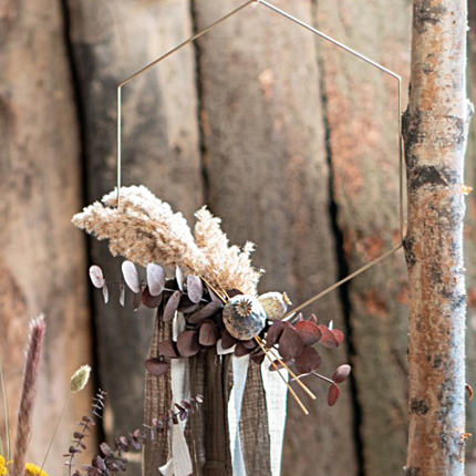 Gouden hanger in de vorm van een hexagon hangt boven een bosje droogbloemen naast een dunne boomstam