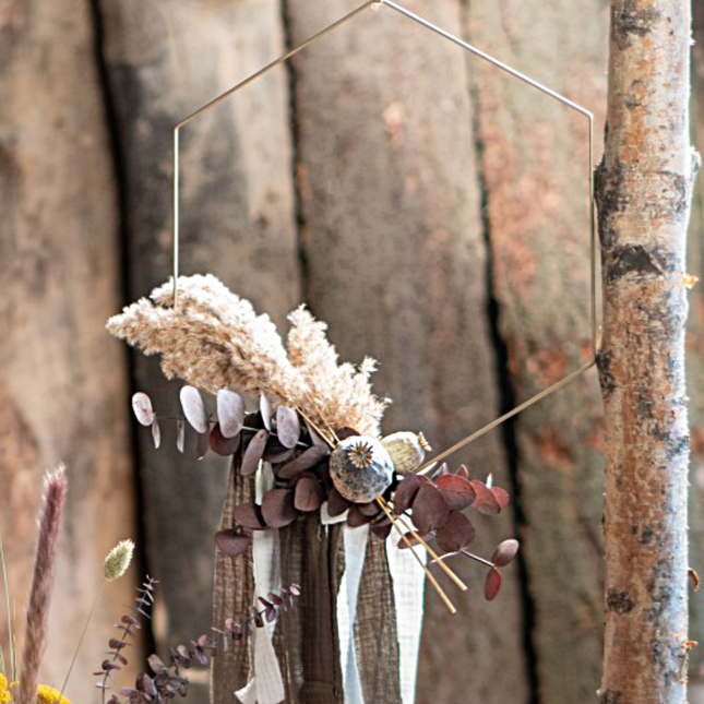 Gouden hanger in de vorm van een hexagon hangt boven een bosje droogbloemen naast een dunne boomstam