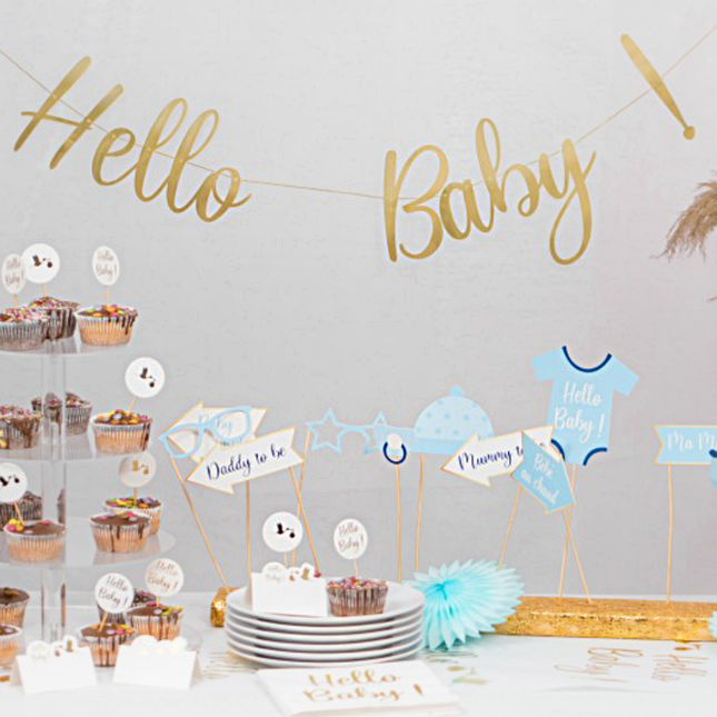Gouden hello baby slinger hangt voor een grijze muur boven een tafel met versiering voor een babyshower