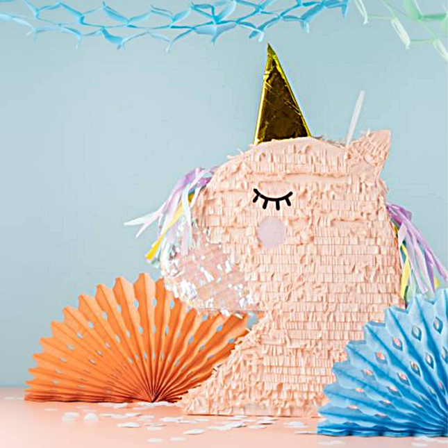 Pinata in de vorm van een roze eenhoorn met gouden hoorn en iridescent effect staat voor een blauwe muur voor een oranje en blauwe waaier