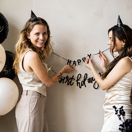 twee vrouwen met zwarte feesthoedjes hangen een zwarte happy birthday slinger op