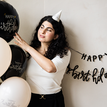 een vrouw met zwarte krullen staat naast creme en zwarte ballonnen en met een creme feesthoedje op