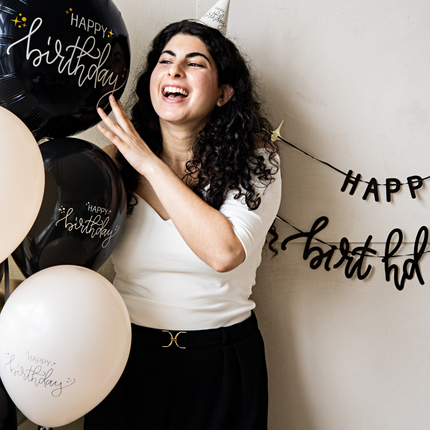 een vrouw met zwarte krullen staat naast creme en zwarte ballonnen en met een creme feesthoedje op
