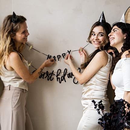 drie vrouwen met zwarte feesthoedjes hangen een zwarte happy birthday slinger op