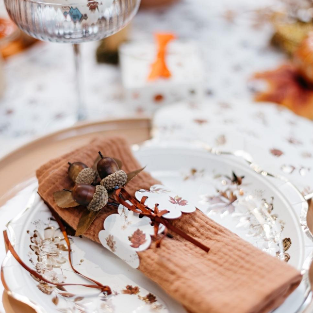 decoratieve takjes met bladeren en eikels liggen op een terracotta servet met een beige servetring met herfstbladeren en wijnrood satijnlint