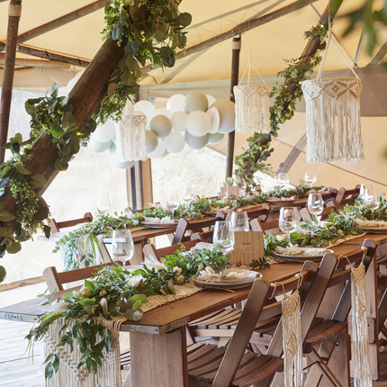 Houten tafel met macrame versiering aan de stoelen en macrame kroonluchters staat onder een tent versierd met groene bladslingers