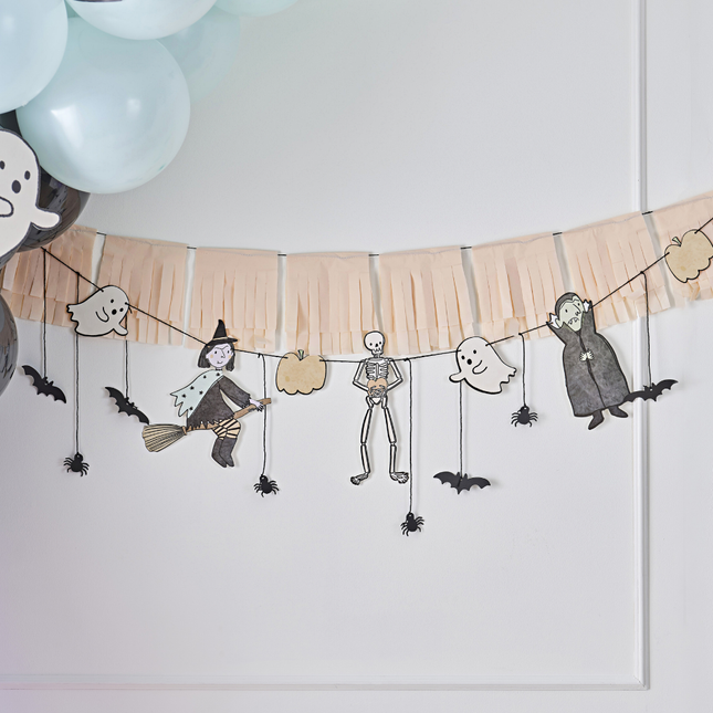 Oranje tasselslinger met spookfiguurtjes zoals een spookje, heks, vampier en skelet hangt voor een witte muur naast een mint ballonnenboog