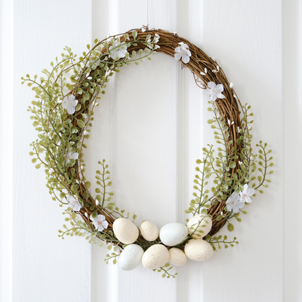 Deurkrans hangt aan een witte deur en is versierd met witte bloemen en paaseieren