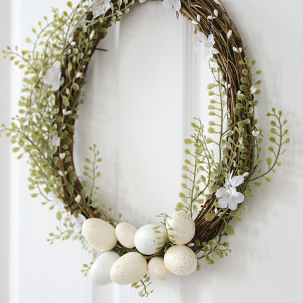 Deurkrans hangt aan een witte deur en is versierd met witte bloemen en paaseieren