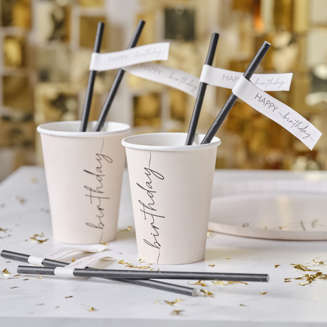 Zwarte rietjes met een nude papiertje eraan met de tekst 'happy birthday' zitten in een nude papieren bekertje met de zwarte tekst birthday erop
