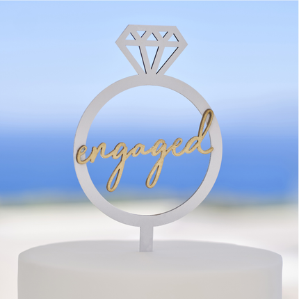 Witte taarttopper in de vorm van een ring met gouden woord engaged zit in een witte taart en staat voor een blauwe achtergrond