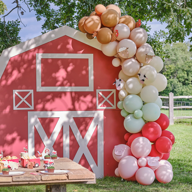 Boerderij met een ballonnenboog in het rood, groen, roze en bruin staat op een grasveld onder een boom achter een houten picknicktafel