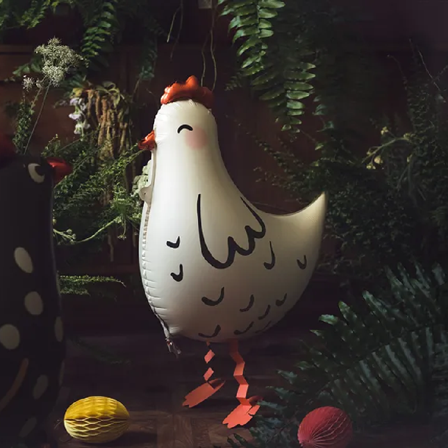 Witte folieballon in de vorm van een kip staat in een zwarte ruimte met palmbladeren