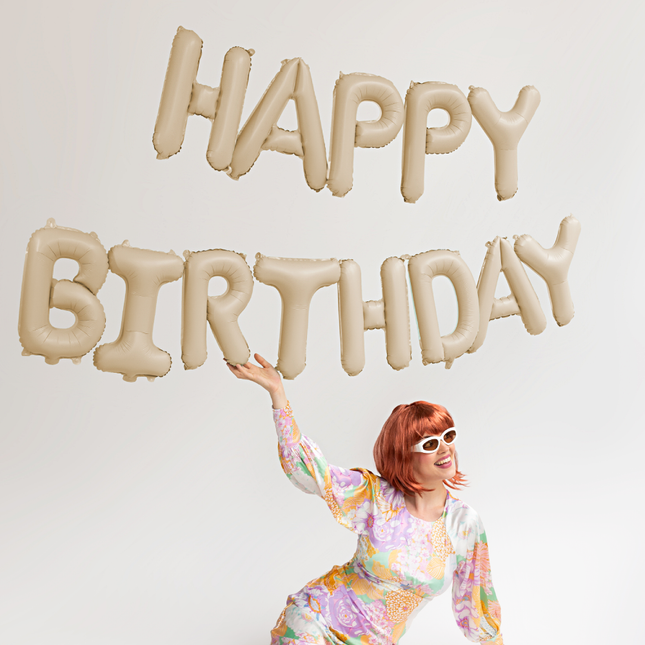 beige ballonnen met de tekst happy birthday hangen boven een vrouw met rood haar