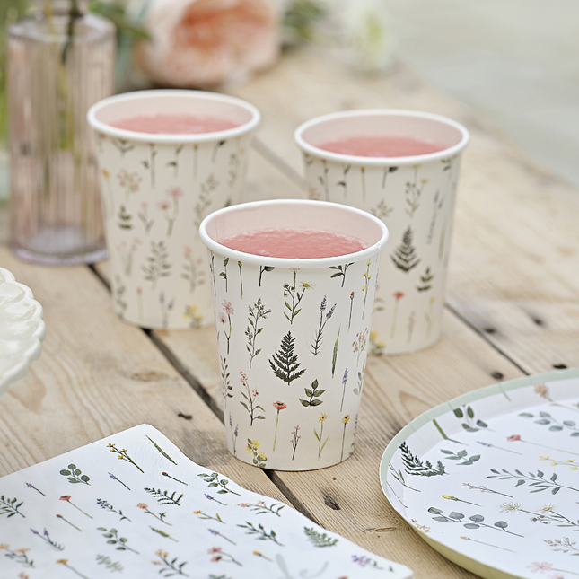Bekers versierd met bloemen en planten staan op een houten tafel naast bijpassende borden en servetten