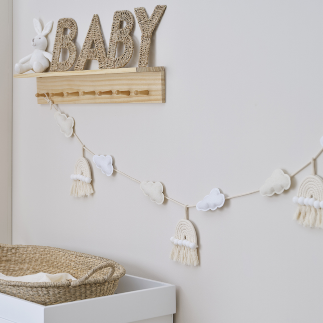 Macrame slinger met wolken en regenbogen hangt in een babykamer met een rieten mand en wit dekentje en rieten letters baby