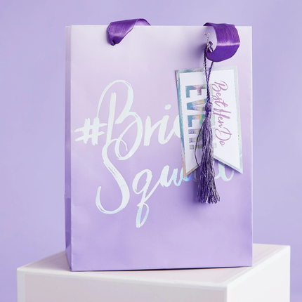 Paars tasje staat op wit blok voor een paarse achtergrond en heeft de iridescent tekst bride squad