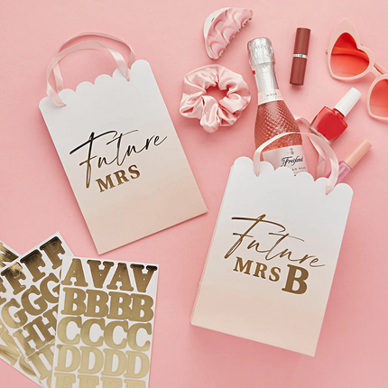 perzikkleurige en witte cadeautasjes met ombre effect en gouden tekst future mrs liggen op een roze tafel en zijn gevuld met nagellak, lippenstift en een zonnebril