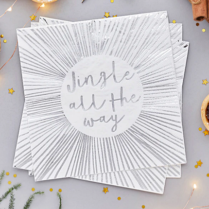 Witte servetten met zilveren strepen en de tekst jingle all the way liggen naast gouden confetti en groene takjes van een dennenboom