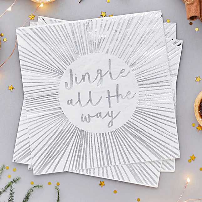 Witte servetten met zilveren strepen en de tekst jingle all the way liggen naast gouden confetti en groene takjes van een dennenboom
