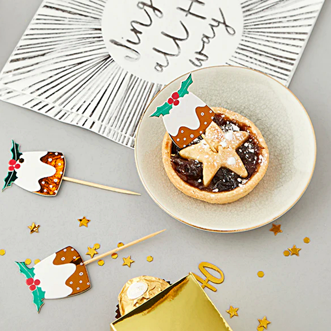 Witte tafel gevuld met kerstversiering, zoals prikkers met een pudding en hulst en een zilveren servet