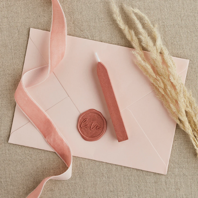 zachtroze envelop is gesloten met rose gouden wax en ligt op een jute tafelkleed naast een roze lint en graan