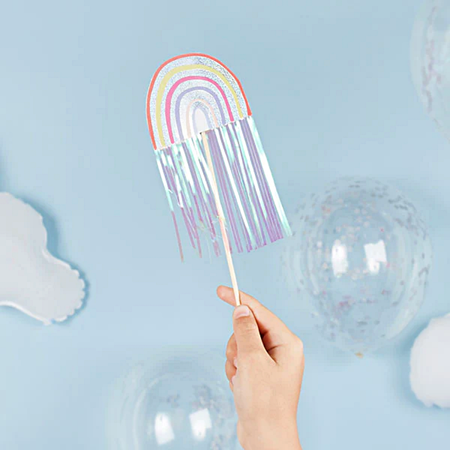 Toverstokje met regenboog en iridescent tassels wordt vastgehouden voor een lichtblauwe achtergrond naast confettiballonnen