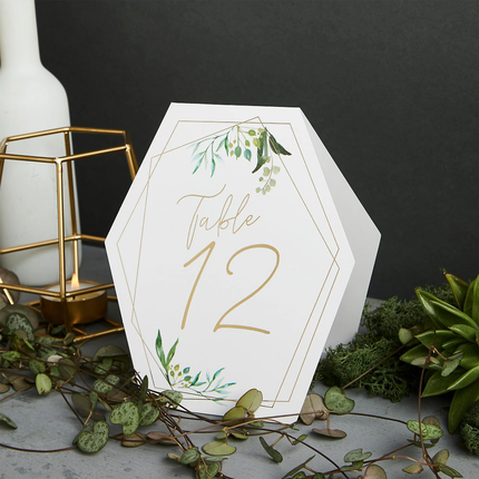 Tafelkaartjes met gouden tekst, groene bladeren en een geometrische vorm staan voor een zwarte muur naast een gouden kaarshouder
