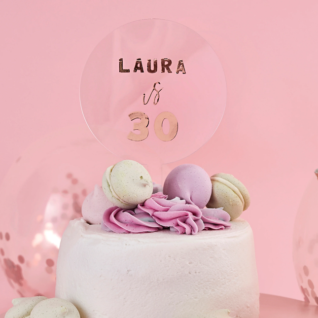taart topper met rose gouden tekst zit in een witte taart versierd met groene en paarse macarons