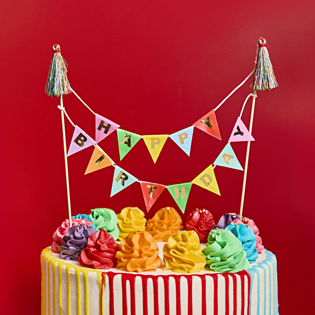 mini slinger voor bovenop een taart met de tekst happy birthday zit in een regenboog drip taart voor een rode achtergrond