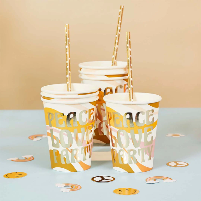 Bekers staan op een blauwe tafel naast confetti en zijn bedrukt met de gouden tekst peace love party en een mosterdgeel en roze patroon