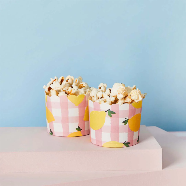 Kartonnen bakjes met roze geblokt patroon en gele citroenen zijn gevuld met popcorn en staan voor een lichtblauwe muur