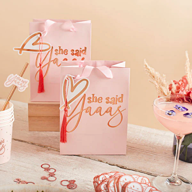 Roze cadeautasje met tassel en hartje en rose gouden tekst she said yaaas staan op wit hout naast een glas met roze gin en paarse bloemetjes
