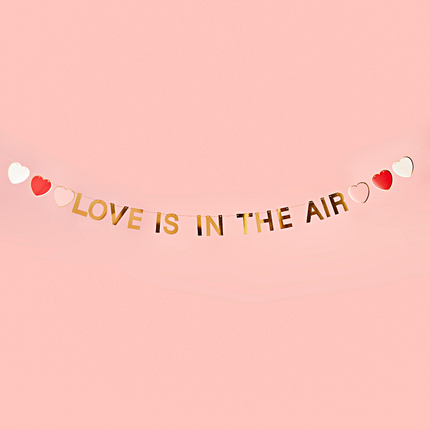 Gouden slinger met de tekst love is in the air en hartjes in het wit, roze en rood hangt voor een lichtroze muur