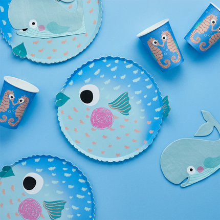 papieren bordje in de vorm van een kogelvis in het blauw met ombre effect ligt op een blauwe achtergrond naast walvis servetten en bekers met zeepaardjes