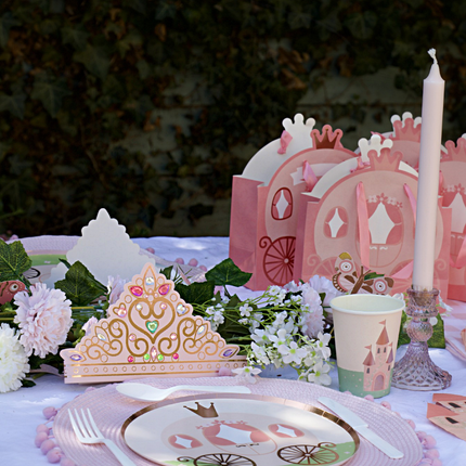 Tafel versierd met een roze tiara, bordjes met een koets erop en cadeautasjes in de vorm van een koets naast een bloemenslinger en een lange kaars