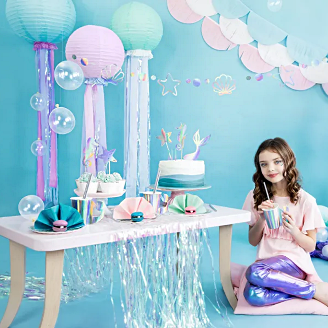 meisje met bruin haar zit voor een blauwe achtergrond naast een versierde tafel met lichtroze en iridescent versiering