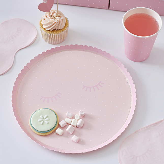 Roze bordje met wimpers en witte stippen en geribbelde rand staan naast een bijpassende, roze beker met servetten in de vorm van een oogmasker