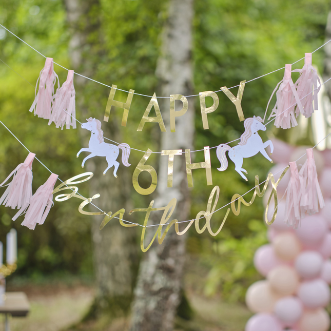 Personaliseerbare slinger met gouden letters, witte paarden en roze franjes hangt in een bos voor een zachtroze ballonnenboog