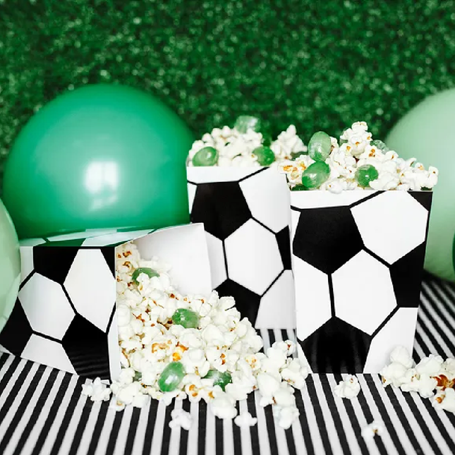 Popcornbakjes met voetbalprint zijn gevuld met popcorn en groene snoepjes en staan op een wit met zwart gestreepte ondergrond