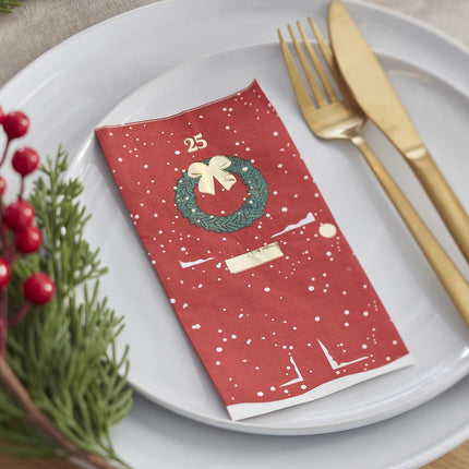 Servet in de vorm van een rode deur versierd voor kerst op een bordje met gouden bestek
