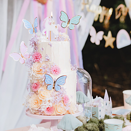 verjaardagstaart versierd met bloemen, pastel vlinders en een eenhoorn kaars staat voor een pastel slinger met vlinders en sterren en een pastel regenboog doek