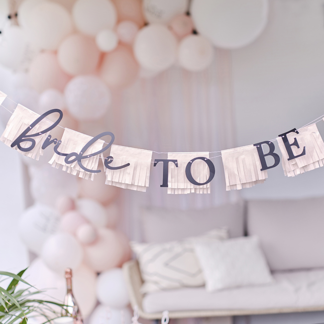 Slinger met roze franjes en de zwarte tekst bride to be hangt voor een roze ballonnenboog en een bank met beige kussens