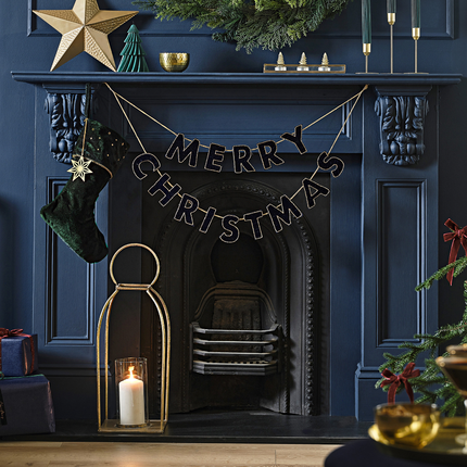 Zwarte velvet slinger met de tekst merry christmas met gouden details hangt voor een donkerblauwe openhaard boven een goud windlicht