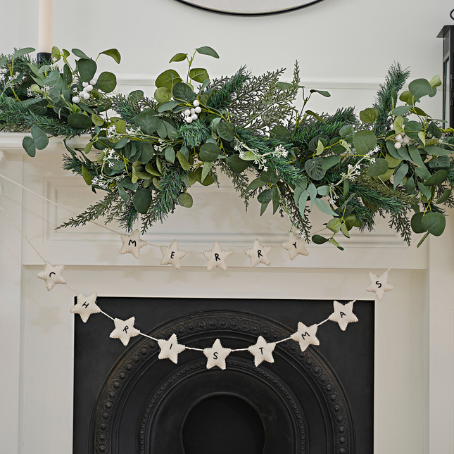 Witte slinger van vilt met sterren en de tekst merry christmas hangt voor een wit met zwarte schouw onder groen gebladerte