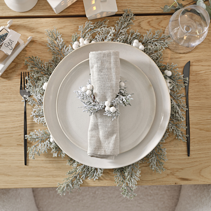 Placemat met groene takjes met sneeuw en witte bessen ligt onder een wit bord met een stoffen servet op een houten tafel