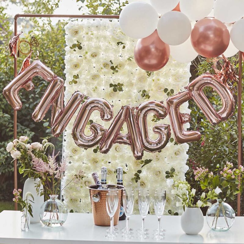 Tafel met champagne en grote folieballon met het woord engaged en een achtergrond van witte bloemen