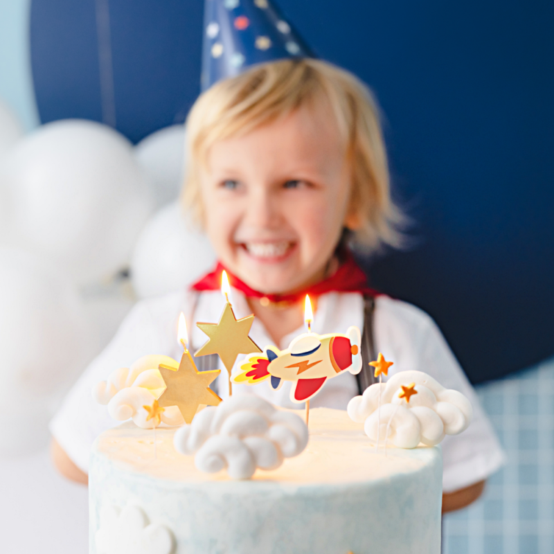 Taart kaarsjes in de vorm van gouden sterren en een vliegtuig worden vastgehouden door ee jongen met een blauw feesthoedje op