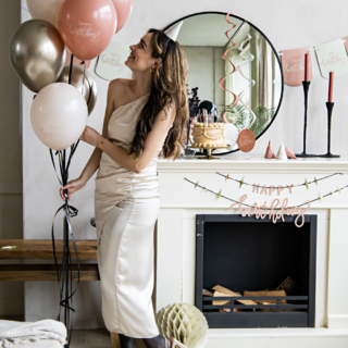 vrouw met een witte jurk houdt een tros roze en gouden ballonnen vast en staat voor een versierde schouw